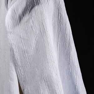 May include: Close-up of a white fabric with a textured, vertical ribbed pattern. The fabric appears to be a lightweight material, possibly linen or cotton, with a subtle sheen. The background is a solid black.
