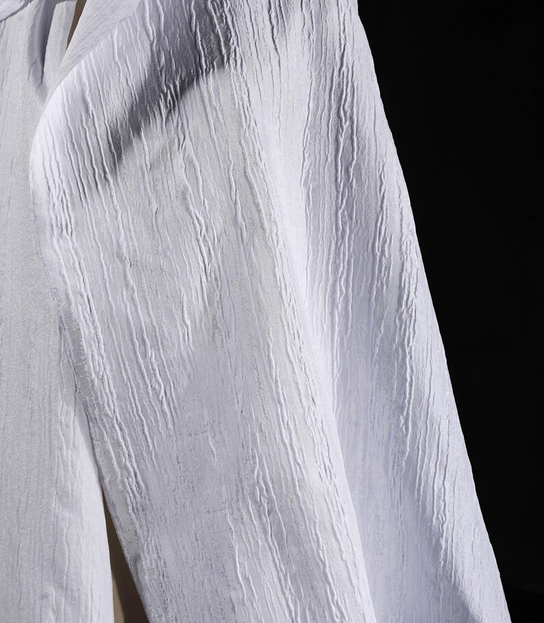 May include: Close-up of a white fabric with a textured, vertical ribbed pattern. The fabric appears to be a lightweight material, possibly linen or cotton, with a subtle sheen. The background is a solid black.