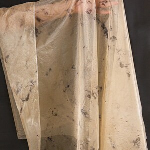 May include: A sheer, beige fabric with dark, irregular splotches. The fabric is draped, revealing the shape of a hand underneath. The material appears lightweight and translucent, with a slightly wrinkled texture. The background is a solid black.