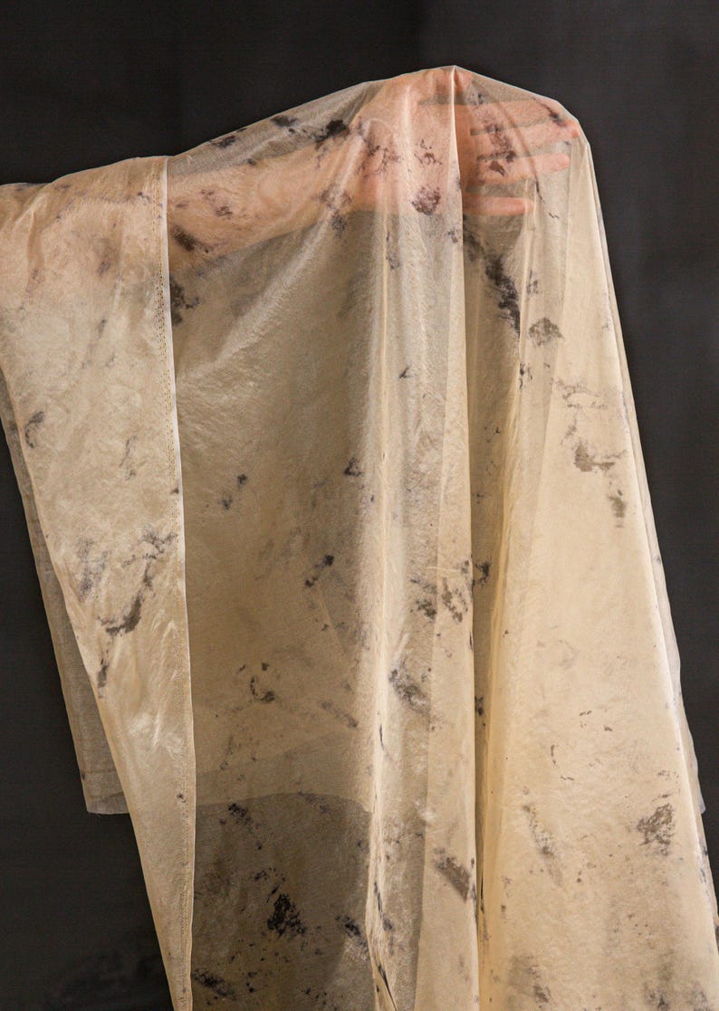 May include: A sheer, beige fabric with dark, irregular splotches. The fabric is draped, revealing the shape of a hand underneath. The material appears lightweight and translucent, with a slightly wrinkled texture. The background is a solid black.