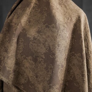 May include: A draped fabric with a textured, mottled pattern in shades of brown and tan. The fabric has a rough, organic appearance, with a visible fringe along one edge. The overall effect is earthy and rustic.