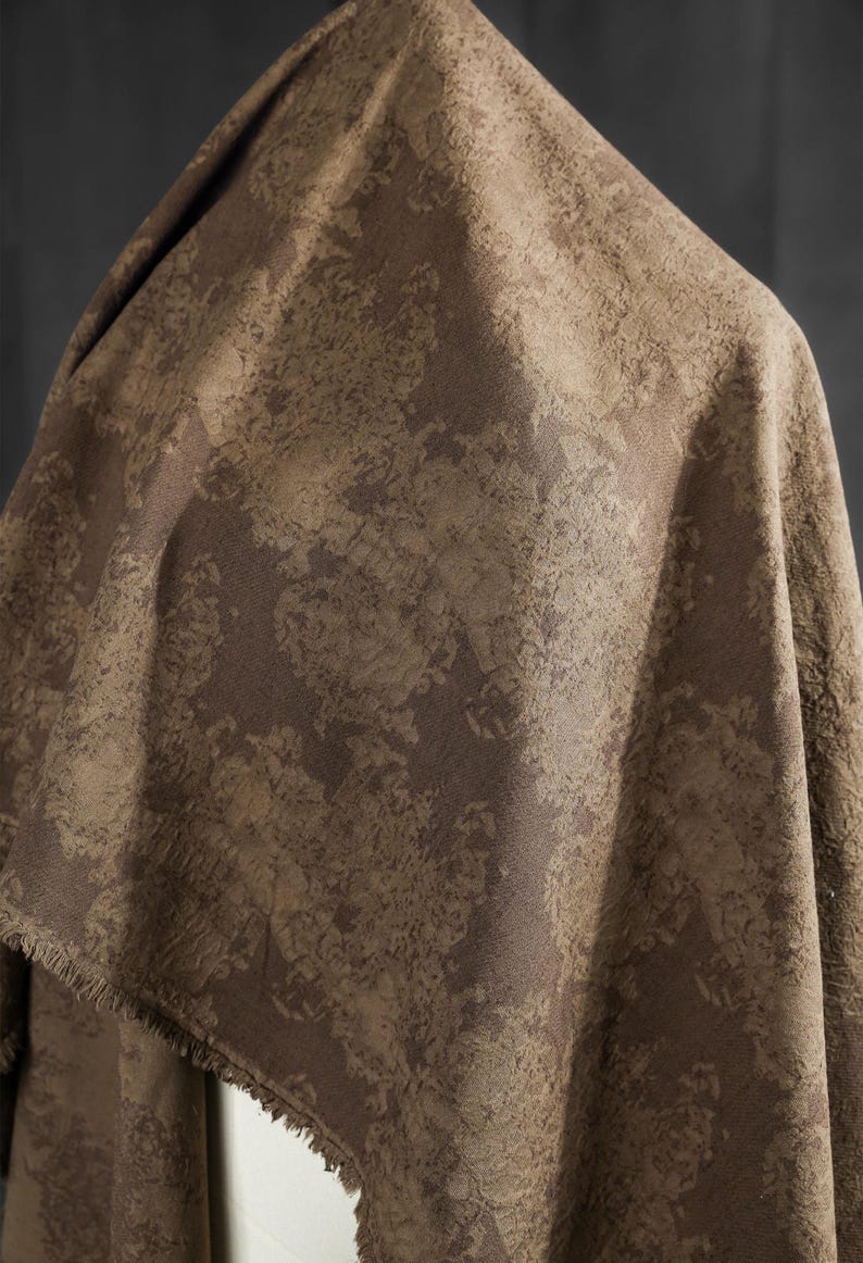 May include: A draped fabric with a textured, mottled pattern in shades of brown and tan. The fabric has a rough, organic appearance, with a visible fringe along one edge. The overall effect is earthy and rustic.