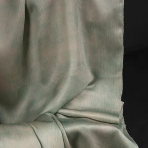 May include: A close-up of draped fabric in shades of pale green and white. The material has a subtle sheen and a soft, flowing texture. The fabric is folded, creating shadows and highlighting the different tones.