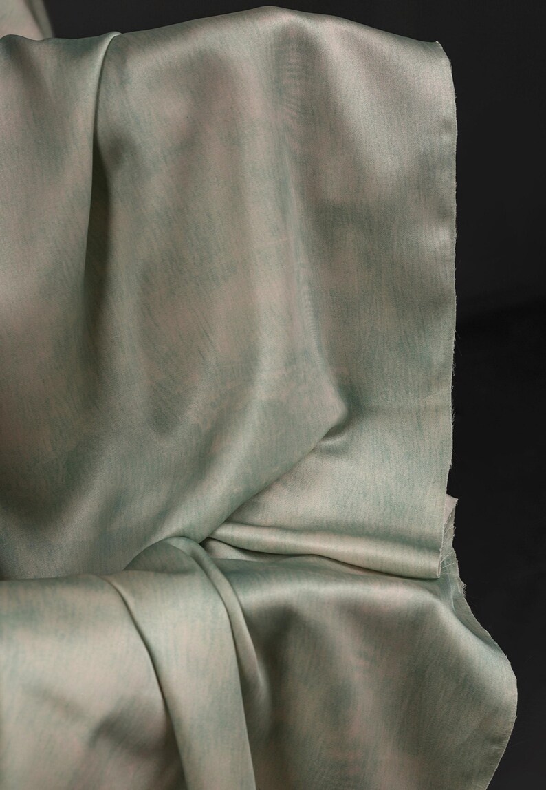 May include: A close-up of draped fabric in shades of pale green and white. The material has a subtle sheen and a soft, flowing texture. The fabric is folded, creating shadows and highlighting the different tones.