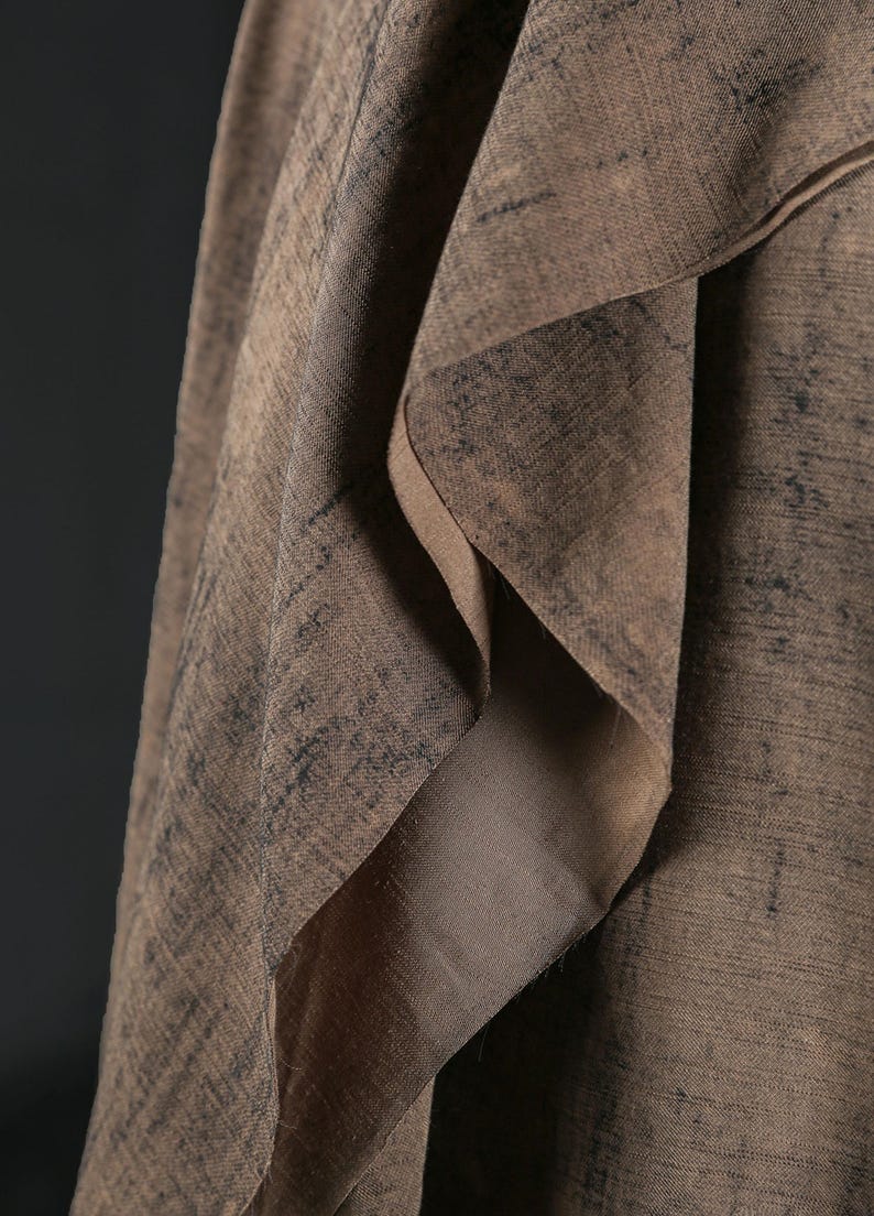 Puede incluir: Primer plano de una tela marr&oacute;n drapeada con textura. El material presenta pliegues en capas y un brillo sutil. Los patrones m&aacute;s oscuros y moteados crean profundidad, sugiriendo un textil de fibra natural. El dise&ntilde;o es intrincado y atractivo.