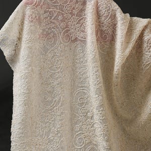 May include: Close-up of a cream-colored lace fabric with an intricate floral and swirl pattern. The fabric is draped, showing the texture and design details. The lace appears to be a delicate material.