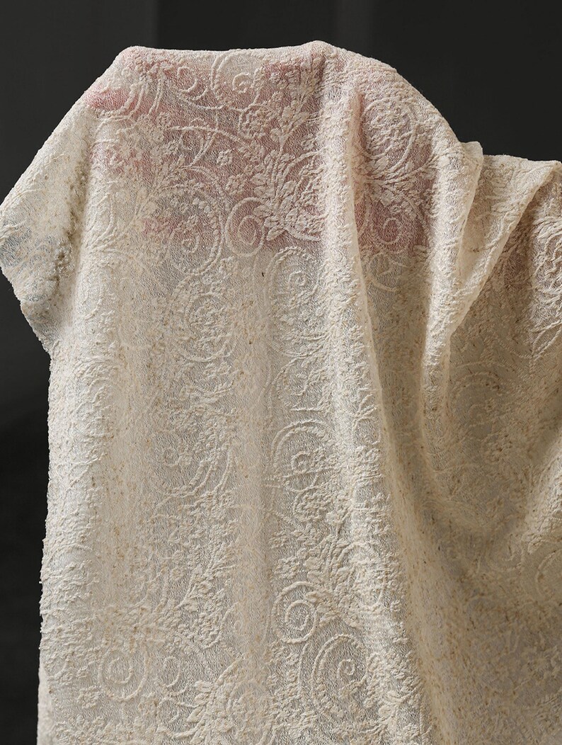 May include: Close-up of a cream-colored lace fabric with an intricate floral and swirl pattern. The fabric is draped, showing the texture and design details. The lace appears to be a delicate material.