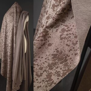 May include: A draped fabric with a speckled pattern in shades of brown and beige. The fabric is draped over a form, showcasing its texture and flow. The design features irregular, darker brown spots against a lighter background, creating a visual contrast.