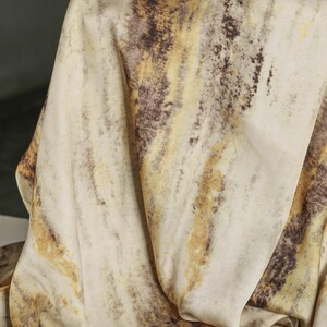 May include: Close-up of a draped fabric with a marbled pattern. The fabric features a cream-colored base with streaks of brown and yellow, creating a natural, organic aesthetic. The folds and shadows add depth.