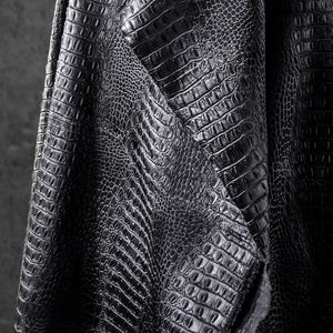 May include: Close-up of black faux leather fabric with a crocodile skin texture. The material is folded, revealing the detailed pattern and texture of the embossed design. The fabric has a matte finish.