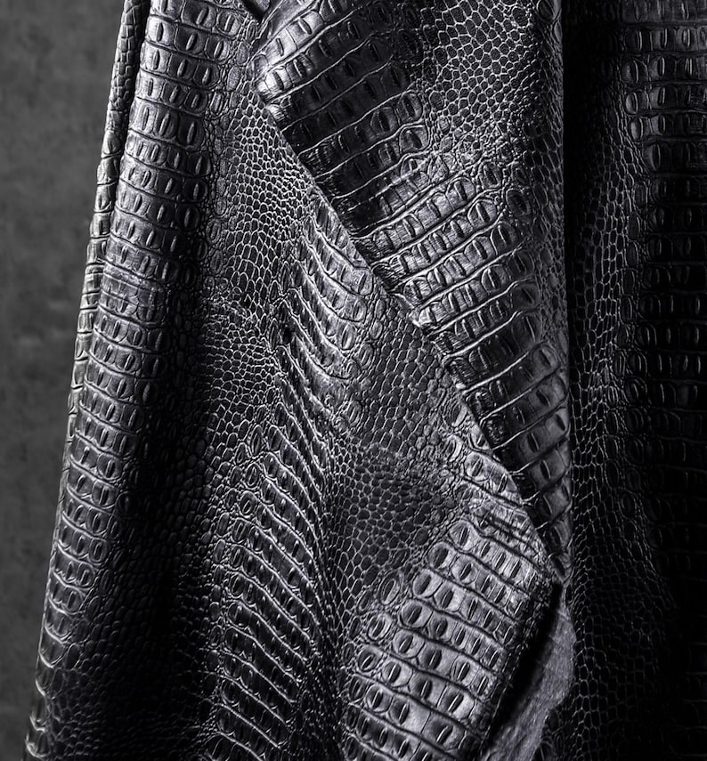 May include: Close-up of black faux leather fabric with a crocodile skin texture. The material is folded, revealing the detailed pattern and texture of the embossed design. The fabric has a matte finish.
