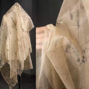May include: Sheer, cream-colored fabric with dark gray mottled patterns. The fabric is draped over a mannequin, showcasing its flowing texture. The image also shows a close-up of the fabric, highlighting its delicate nature and design.