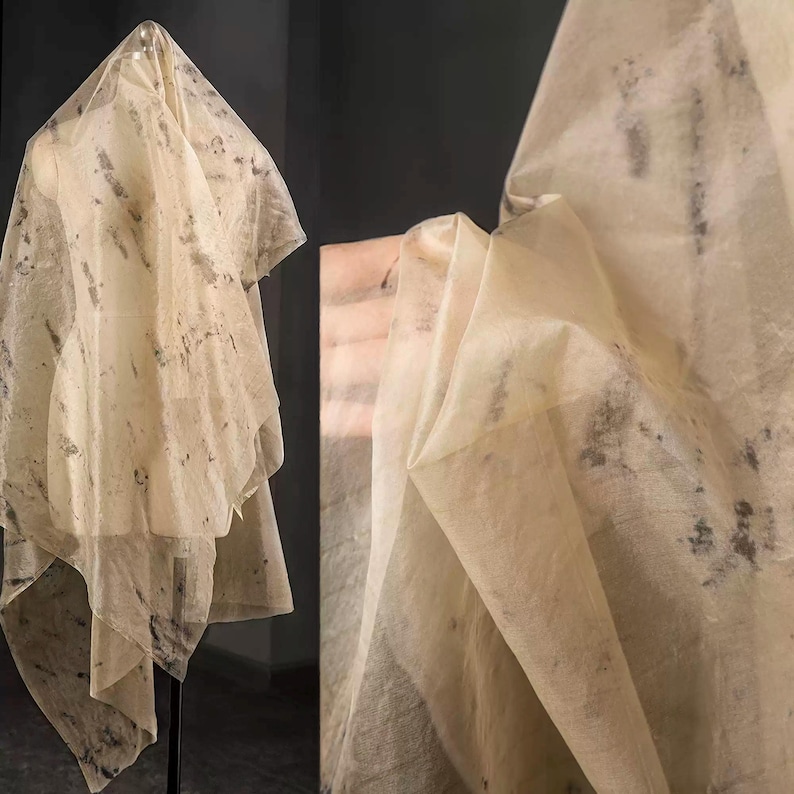 May include: Sheer, cream-colored fabric with dark gray mottled patterns. The fabric is draped over a mannequin, showcasing its flowing texture. The image also shows a close-up of the fabric, highlighting its delicate nature and design.