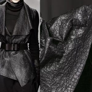 May include: A black leather vest with a textured, crocodile-like pattern. The vest has a draped front and is cinched at the waist with a wide black belt. The outfit includes a black turtleneck and long-sleeved top.