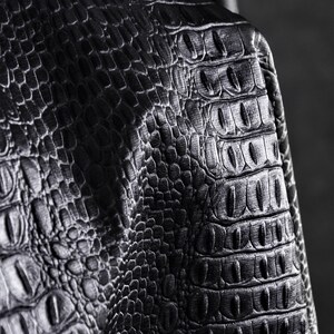 May include: Close-up of black leather with a crocodile skin texture. The leather has a detailed pattern of scales and ridges, creating a realistic and textured appearance. The lighting highlights the depth and dimension of the material.