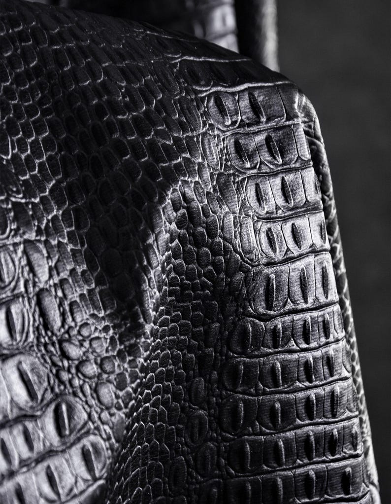 May include: Close-up of black leather with a crocodile skin texture. The leather has a detailed pattern of scales and ridges, creating a realistic and textured appearance. The lighting highlights the depth and dimension of the material.