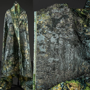 Puede incluir: Una tela lujosa con un patrón de bosque en tonos verdes, dorados y negros. La tela está drapeada sobre un maniquí, mostrando su textura y diseño. El borde de la tela es visible, revelando un ribete con flecos.