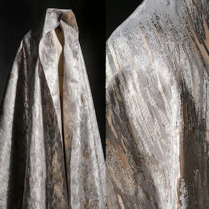 May include: A draped fabric with a metallic sheen, featuring a blend of silver, gray, and gold tones. The material has a textured, vertical stripe pattern, creating a luxurious and elegant appearance. The fabric is draped over a form, showcasing its flow and drape.