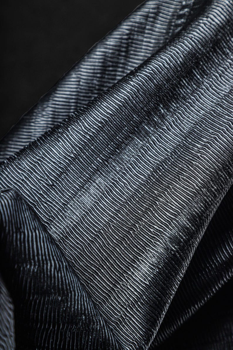 May include: Close-up of a textured fabric with a pattern of parallel, slightly curved lines. The fabric appears to be a dark gray or black color, with subtle highlights creating a sense of depth and dimension. The material is folded.
