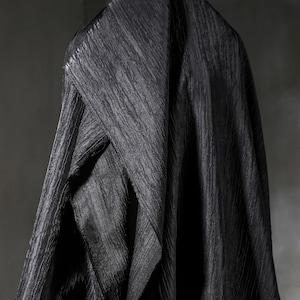 May include: A draped, textured black fabric with a diagonal ribbed pattern. The fabric is folded and hangs in soft, flowing lines, creating shadows and highlights. The overall effect is one of luxury and depth.
