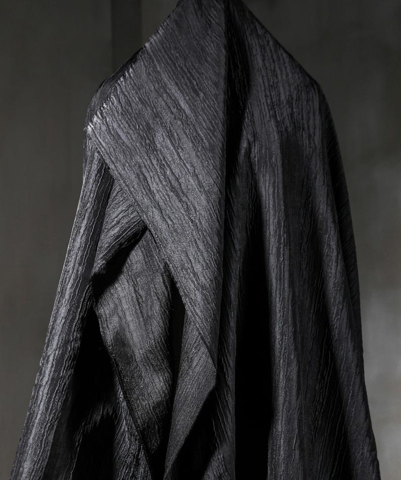May include: A draped, textured black fabric with a diagonal ribbed pattern. The fabric is folded and hangs in soft, flowing lines, creating shadows and highlights. The overall effect is one of luxury and depth.