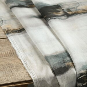 May include: White fabric with an abstract watercolour design in shades of grey, black, and brown. The fabric is draped over a wooden surface, showing its texture and pattern. The edges are unfinished.