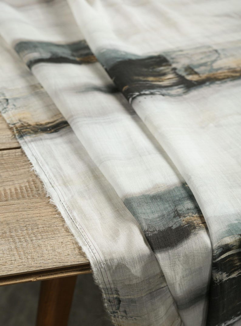 May include: White fabric with an abstract watercolour design in shades of grey, black, and brown. The fabric is draped over a wooden surface, showing its texture and pattern. The edges are unfinished.
