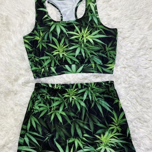 Op de afbeelding: Een tweedelige outfit met een groene en zwarte marihuana-bladprint. Het bovenstuk is een tanktop met een ronde hals en het onderstuk is een short.