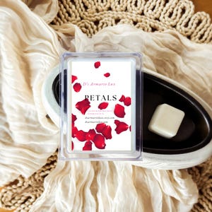 May include: A clear plastic package containing wax melts with a label that reads "Petals" and features red rose petals. A white wax melt cube sits in a black and white ceramic warmer. The background includes a beige fabric and woven material.