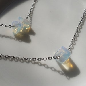 May include: A silver chain necklace with two iridescent opal stones. The necklace is displayed on a white ceramic bowl.