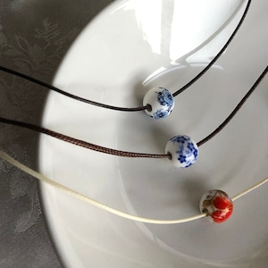 May include: Three necklaces with ceramic beads. One bead is red with white detailing, and two are white with blue floral patterns. The cords are brown, black, and cream, displayed on a white plate.