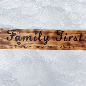 Op de afbeelding: Een houten bord met de woorden "Family First" in zwarte cursieve letters. Het bord heeft ook de woorden "Today, Tomorrow, Always" in kleinere zwarte letters.