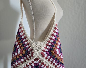 May include: A handmade crochet shoulder bag in a granny square pattern. The bag features a cream-colored strap and trim, with a colorful body of white, burgundy, navy, and mustard squares. The bag is displayed on a mannequin.