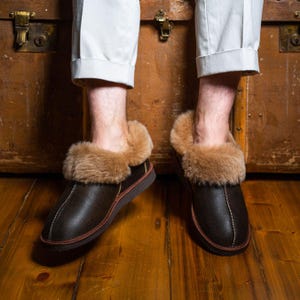Könnte beinhalten: Braune Lederslipper mit Lammfellfutter und brauner Ledersohle. Die Slipper werden von einer Person getragen, die auf einer Holzkiste sitzt.