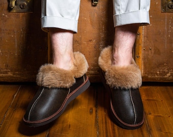 May include: Brown leather slippers with a shearling lining and a brown leather sole. The slippers are worn by a person sitting on a wooden chest.