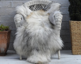 Puede incluir: Una piel de oveja de pelo largo y esponjoso que presenta una mezcla de tonos blancos, grises y marrones claros. La piel está colocada sobre una silla de mimbre blanca e incluye el texto impreso 115 x 80 cm XXL en la parte inferior.