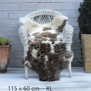 May include: A sheepskin rug featuring a mottled pattern of cream and dark brown fur. The rug is draped over a white wicker chair and includes the text 115 x 60 cm XL at the bottom, indicating its size in centimetres and its extra large classification.