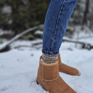 May include: A pair of brown suede ankle boots with a shearling lining. The boots are worn by a person standing in the snow.