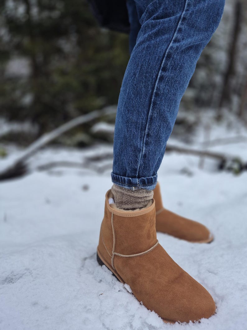 May include: A pair of brown suede ankle boots with a shearling lining. The boots are worn by a person standing in the snow.