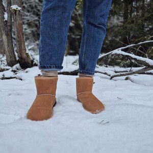 May include: A pair of brown suede boots with a shearling lining, worn by a person standing in the snow. The person is wearing blue jeans.