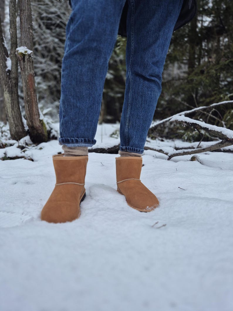 May include: A pair of brown suede boots with a shearling lining, worn by a person standing in the snow. The person is wearing blue jeans.