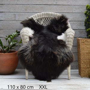 May include: A long-haired, dark brown and black sheepskin rug with white accents draped over a white wicker chair. The rug is labelled as XXL and measures 110 cm by 80 cm, providing a thick, textured appearance with natural colour variations.