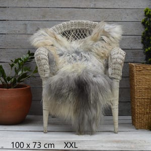 Può includere: Una pelle di pecora a pelo lungo, grigia e beige, drappeggiata su una sedia in vimini bianco. Il tappeto presenta una sfumatura di colore naturale e misura 100 x 73 cm, etichettato come XXL. La consistenza è folta, ispida e voluminosa.