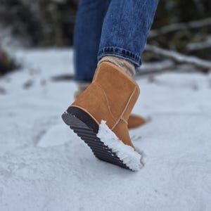 May include: A close-up of a brown suede winter boot with a black rubber sole. The boot is covered in snow and is being worn by a person walking in the snow.