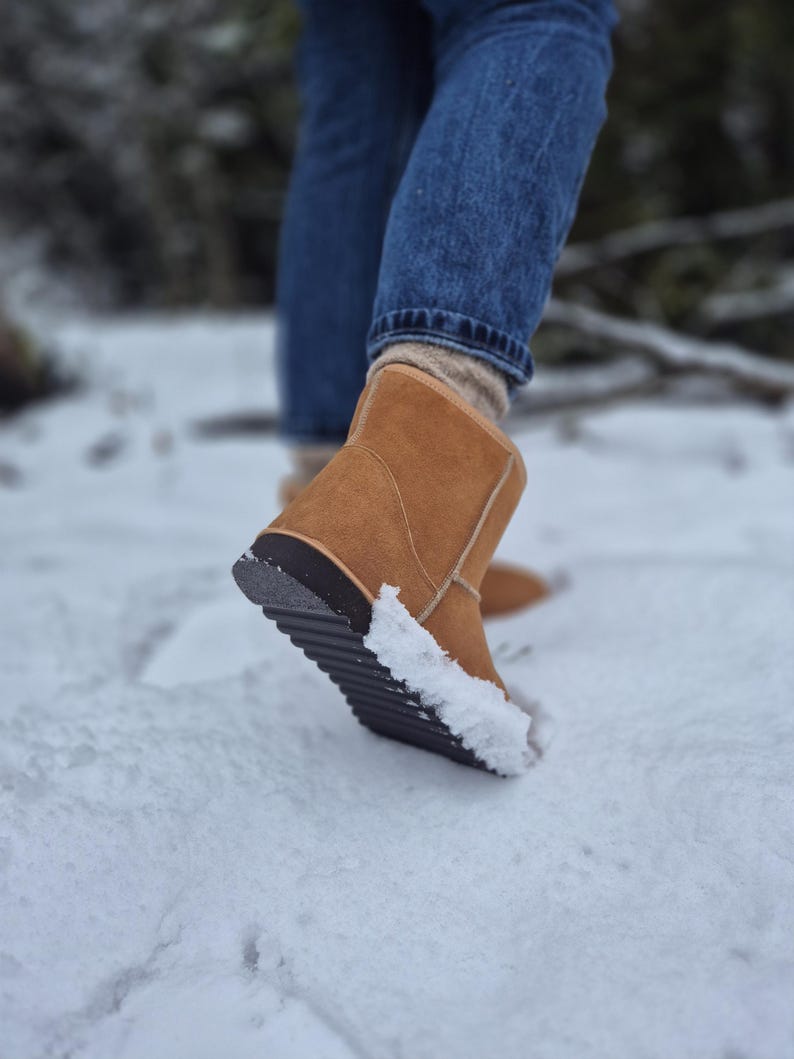 May include: A close-up of a brown suede winter boot with a black rubber sole. The boot is covered in snow and is being worn by a person walking in the snow.