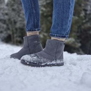 May include: A pair of gray suede boots with a faux fur lining. The boots are worn by a person wearing blue jeans and standing in the snow.