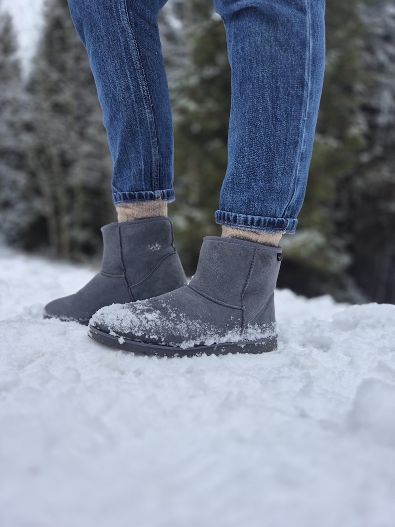 May include: A pair of gray suede boots with a faux fur lining. The boots are worn by a person wearing blue jeans and standing in the snow.