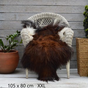 May include: A long-haired sheepskin rug featuring a deep brown center with white and grey accents along the edges. The rug is draped over a white wicker chair and includes the printed text 105 x 80 cm XXL at the bottom of the image.
