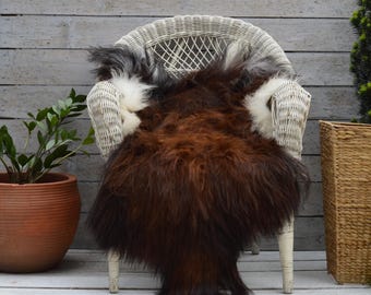 May include: A long-haired sheepskin rug featuring a deep brown centre with white and grey accents along the edges. The rug is draped over a white wicker chair and includes the printed text 105 x 80 cm XXL at the bottom of the image.