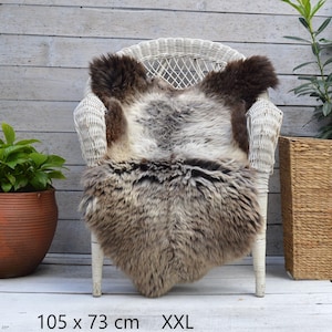 Puede incluir: Una alfombra de piel de oveja esponjosa con una mezcla de pelaje marrón oscuro y gris claro, colocada sobre una silla de mimbre blanca. La alfombra está etiquetada como XXL y mide 105 cm por 73 cm, con un pelo grueso y texturizado.